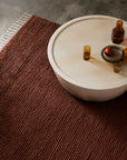 A round white coffee table with amber glass bottles and a bowl with an apple sits on the Armadillo&Co Malawi Rug - Amber, bringing an artisanal feel to the concrete floor.