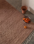 The Malawi Rug - Blush by Armadillo&Co lies on a speckled floor, with a gray plate of persimmons, a single persimmon beside it, and an empty terracotta cup placed near a white wall.