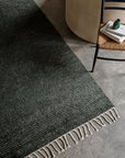 The Malawi Rug - Kelp by Armadillo&Co, a dark green hand-knotted wool piece, crafted by Fair Trade artisans, rests on a concrete floor beside a woven chair and a small table with books and a green decor item.