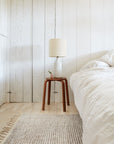 A cozy bedroom with white wooden walls, a bed with white bedding, a small brown side table with a white textured lamp and plant, and an artisanal touch from the Malawi Rug - Oatmeal by Armadillo&Co on the floor.