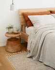 Minimalist bedroom featuring the Air Bed by Ethnicraft in European oak, accented with beige and rust pillows, light gray bedding, and a small wooden side table displaying plants. A neutral textured rug lies on the light wood floor, and a pendant light hangs above the table.