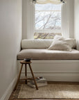 A cozy window nook features a cushioned bench, pillow, throw blanket, and small wooden stool with books and a bowl. Sunlight highlights the artisanal charm of the Armadillo&Co Malawi Rug in Oatmeal beneath this inviting space.