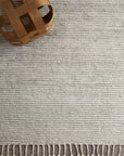 The Malawi Rug - Oatmeal by Armadillo&Co, a hand-knotted wool rug in light gray with white tassels, adds artisanal style on a concrete floor. A wooden woven basket is visible in the top left corner.
