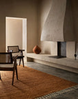 A minimalist living room features textured beige walls, a sculptural fireplace, wooden chairs with cane backs, and the Malawi Rug - Saffron by Armadillo&Co. A round terracotta vase on a built-in ledge enhances the cozy, artisanal atmosphere.