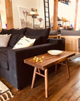 A cozy living room with a dark sofa, neutral pillows, and the SOBU Mars Live Edge Bench in walnut adds natural style. Wooden floors and beams enrich the space, accented by modern furniture and a black ladder in the background.
