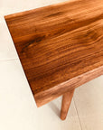 Close-up of the Mars Live Edge Bench - Walnut by SOBU, showcasing natural American walnut grain and a smooth finish. The benchs leg is visible against a light-colored floor.