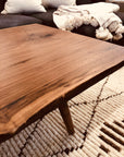A close-up of the SOBU Mars Live Edge Coffee Table in walnut sits on a textured cream and brown rug, beside a dark sofa with neutral pillows and a white tasseled throw blanket.