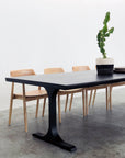 The SOBU Mars Live Edge Dining Table in Black Ash with Black T Legs is paired with light wooden chairs, featuring a potted cactus centerpiece. The setting is against a plain white wall and concrete floor.