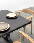The SOBU Mars Live Edge Dining Table in Black Ash with Black T Legs is paired with two rustic ceramic plates and light wooden chairs with curved backs, set on a concrete floor in a minimalistic space.