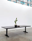 The Mars Live Edge Dining Table - Black Ash with Black T Legs by SOBU stands on a concrete floor, accented by a potted cactus and books. A white wall and large industrial windows complete the minimalist scene from the refined Mars Collection.