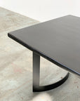 A close-up of the SOBU Mars Live Edge Dining Table, featuring a black ash rectangular top and curved geometric black metal legs, displayed on a light, worn concrete floor.
