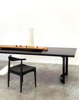 A minimalist Mars Live Edge Dining Table in black ash with curved legs by SOBU pairs with a matching chair. On the table sits an American ash tray holding nuts and a glass carafe of water, set against a white wall and concrete floor.