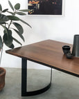 The SOBU DZ TRIAL Mars Live Edge Dining Table with black metal legs stands by a rubber plant in a terracotta pot. On the table are a dark cup, bowl, and tall vase, while a framed photo hangs on the white wall behind.