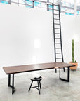 A modern, minimal room features the SOBU DZ TRIAL Mars Live Edge Dining Table, paired with a black stool, a tall ladder against a white wall, and a large potted cactus. Ample light and windows give the space a clean, airy vibe.