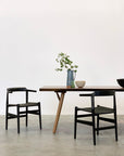 A minimal dining setup from SOBUs Mars Live Edge Dining Table in walnut with wood legs includes two black chairs, a ceramic bowl, a black bowl, and a dark pitcher with green leafy branches on a concrete floor against a plain white wall.