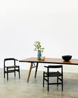 A minimalist dining setup featuring the SOBU Mars Live Edge Dining Table in walnut with wood legs, two black chairs, a black bowl, a white bowl, and a tall vase with green branches against a white wall and concrete floor.