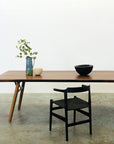 The SOBU Mars Live Edge Dining Table in walnut stands with wood legs, topped with a black bowl, light bowls, and a blue jug of green branches. A black chair faces it on concrete flooring against a plain white wall.