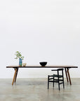 A minimalist scene from SOBU’s Mars Collection features the Mars Live Edge Dining Table in Walnut with Wood Legs, paired with a black chair, a green vase with branches, and two bowls against a plain white wall and concrete floor.