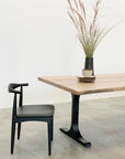 The Mars Live Edge Dining Table in White Ash with Black T Legs by SOBU features stacked plates and a black vase with tall grass stems, paired with a curved-back black chair on a concrete floor.