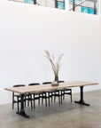 The SOBU Mars Live Edge Dining Table in white ash with black T legs is surrounded by six black chairs. A tall vase with dried grass sits on top. The room has white walls, concrete floors, and large windows above.
