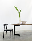 A minimalist scene features a black chair beside the SOBU Mars Live Edge Dining Table in White Ash with Curved Legs. On the table sit a glass vase with tall green leaves, stacked books, and a light bowl against a plain white wall.