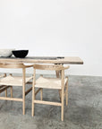 The SOBU Mars Live Edge Dining Table in White Ash with Wood Legs stands on a concrete floor, paired with two light wooden chairs, topped with a decorative runner and bowls, set against a plain white wall.