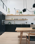 Modern kitchen with black cabinets, open wooden shelves, light wood flooring, and a Mars Live Edge Dining Table – White Ash with Wood Legs by SOBU. Minimalist pendant lighting, white tile backsplash, solid American ash accents, and a high ceiling complete the look.