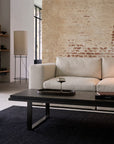 A modern living room featuring the Ethnicraft Mellow Modular Sofa in off white, a black rectangular coffee table, wine glass, book, black rug, standing lamp, and rustic exposed brick wall. Large windows fill the space with natural light.