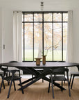 A modern dining room with the Mikado Dining Table Oval in Black Oak by Ethnicraft, six black chairs, a beige rug, two vases with branches, large windows framing trees, white curtains, and a glowing fireplace to the right.