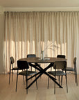 A modern dining area featuring the Ethnicraft Mikado Dining Table Round in black oak, six black chairs, minimalist decor with candles and a vase with branches, and floor-to-ceiling beige curtains in the background.