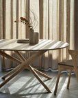 The Ethnicraft Mikado Dining Table Round - Oak, with crisscross legs and elegant design, sits by beige curtains. Sunlight casts shadows as it holds two vases with dried branches beside a wooden chair.