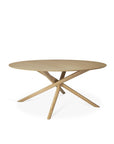 The Mikado Dining Table Round - Oak by Ethnicraft features a smooth European oak top and four intersecting angled legs, showcasing the brand’s modern minimalist design with a light, natural finish.