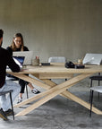 Two people work on laptops at the Ethnicraft Mikado Meeting Table - Oak in a modern office. The solid oak table holds a camera, notebook, pens, and coffee cups, and is surrounded by chairs.