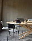 The Mikado Meeting Table - Oak by Ethnicraft features angled legs and is paired with four gray cushioned chairs. On the table are two laptops, a coffee cup, a notebook, and pencils—all set against a neutral gray wall.