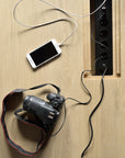 A smartphone and a DSLR camera are connected to a black power strip built into the oak surface of the Ethnicraft Mikado Meeting Table, with charging cables plugged into the outlets.