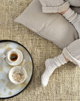 A person in cozy knit socks and loungewear sits on a beige cushion atop a textured carpet, beside an Ethnicraft Mist Gold Organic Glass Tray (Round/Small) holding a cup of tea, a bowl of granola with milk, and a spoon.
