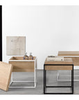 Three Ethnicraft Monolit Side Tables—solid oak tops with black metal frames—are arranged on a gray floor. One table’s top is removed to reveal bottles inside, and a small painting decorates the white wall behind.