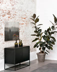 Minimalist room with the Ethnicraft Monolit Sideboard - 2 door, black metal frame, three green glass vases, a painting on a white brick wall, large potted rubber plant, and gray rug on concrete floor.