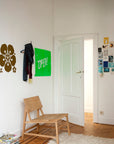 A minimal room featuring the Ethnicraft N2 Lounge Chair in oak on a white rug, with magazines, a floral wall decal, a scarf-draped coat rack, a bright green OPEN! sign, and postcard collage near a white door.
