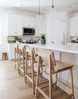 A modern kitchen with white cabinets, a white countertop island, and light wood floors features four Ethnicraft N3 Counter Stools in oak. Pendant lights brighten the island and potted plants bring greenery to the space.