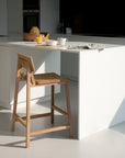 A modern kitchen showcases an Ethnicraft N3 Counter Stool - Oak at a white island, where a tray with a teapot, cups, and fruit sits. Sunlight streams in as a newspaper rests on the countertop, casting gentle shadows.