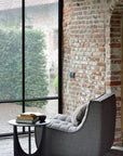 The Ethnicraft N701 Sectional Sofa in dark grey sits beside a small round black table with books and a cup, near large windows overlooking greenery and old brick walls—a perfect spot for fans of European design.