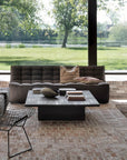 Modern living room with European design features Ethnicrafts N701 Sectional Sofa in Dark Grey, a wire chair, black cabinet, and square coffee table on brick flooring. Large windows overlook a green lawn and riverside trees.