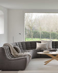 A modern living room features the Ethnicraft N701 Sectional Sofa in Dark Grey, a round wooden coffee table, and a large window with views of leafless trees and greenery. The minimalistic space showcases European design in neutral tones.