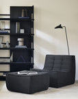 A modern living room with an Ethnicraft N701 Sectional Sofa in graphite high-performance fabric, a matching ottoman holding an open book, a black floor lamp, and a sleek shelving unit with decor set against a beige wall.