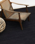 A wooden chair with a woven cane backrest and angled armrests sits on the Nest Rug in Charcoal by Armadillo&Co. A round wicker object is partially visible to the left, creating a modern, minimalist setting.