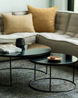 A modern living room features a beige sectional sofa, tan cushions, and Ethnicrafts Nesting Coffee Table Charcoal - Set of 2 with black metal bases holding books and a cup, all atop a textured gray rug for a stylish finishing touch.