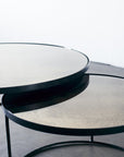 The Ethnicraft Nesting Coffee Table Clear - Set of 2 features two sleek, round tables with slim black metal legs, nested together on a smooth concrete floor by a white wall, the smaller table neatly tucked under the larger.