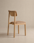 The Noki Dining Chair | Leather by Wooden Stories is a minimalist solid wood chair with a light brown padded leather seat and backrest. Perfect for home or contract use, it’s shown on a neutral background and angled slightly away from the camera.
