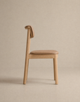 The Noki Dining Chair | Leather by Wooden Stories features a minimalist solid wood design, tan leather seat, and distinctive curved backrest, displayed in profile against a neutral beige background.